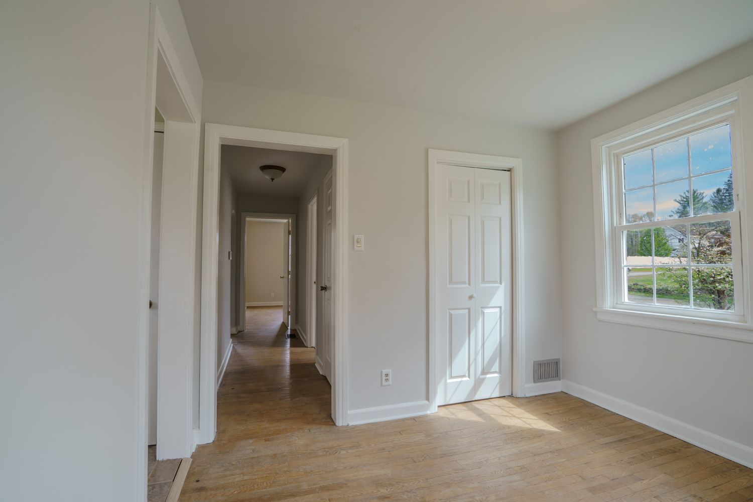 Bright empty bedroom with light wood floors, white walls, closet door, and a window with natural light.