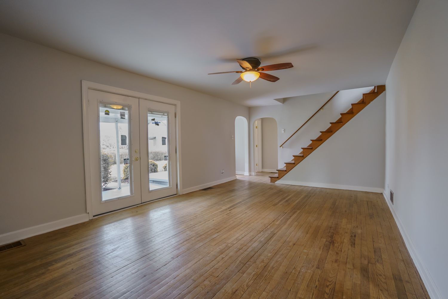 Open living room with hardwood floors, ceiling fan, French doors, arched doorway, and staircase to upper level.
