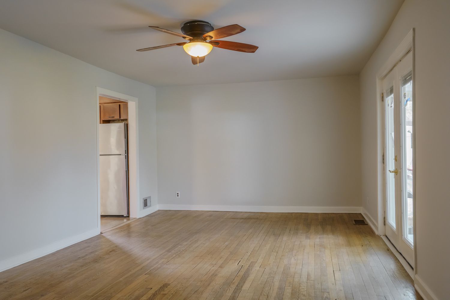 Vacant living room with hardwood floors, ceiling fan, white walls, and glass doors letting in natural light.