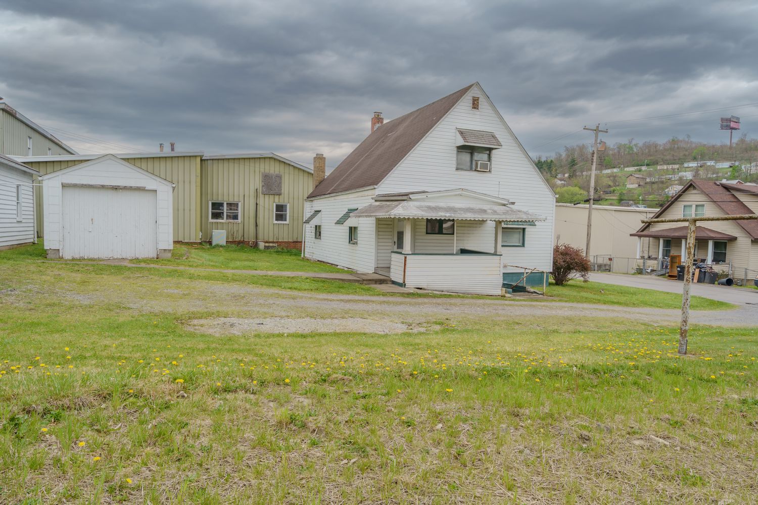 A modest white clapboard house with a detached garage sits on a grassy lot under cloudy skies in a small town.