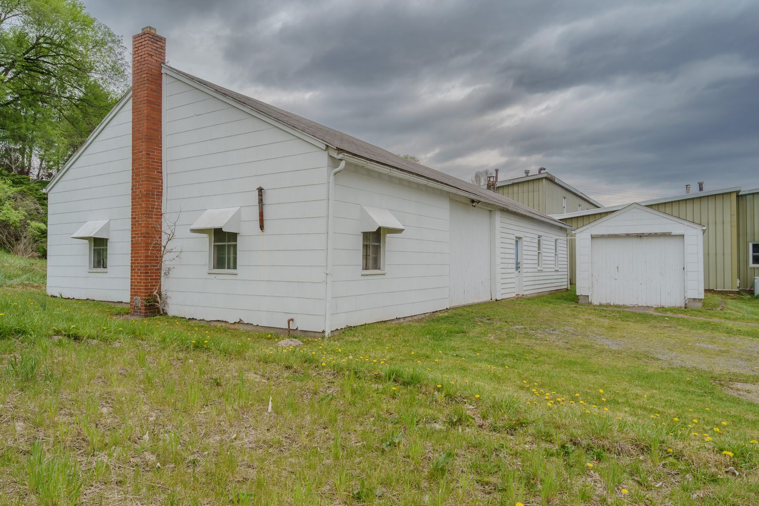 White farmhouse with brick chimney and detached garage on green lawn under cloudy sky.