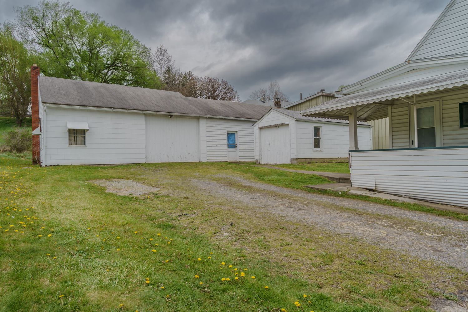 White rural outbuildings with a gravel driveway on a cloudy day, surrounded by green grass and trees.