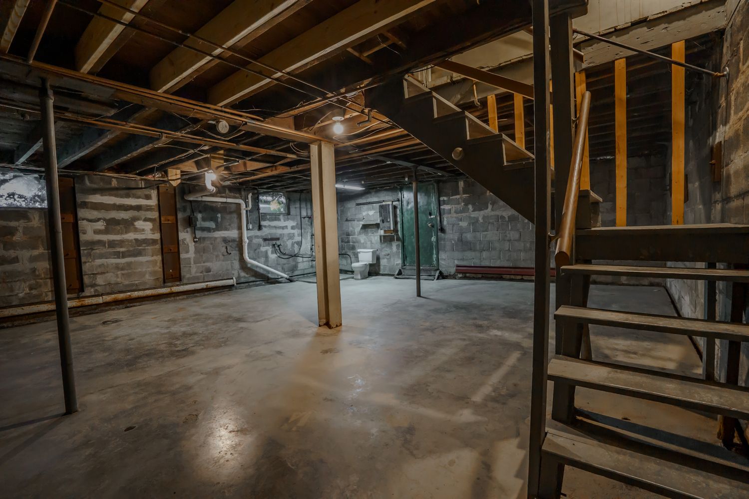 Unfinished basement with exposed ceiling joists, concrete floors, metal support columns, and stairs leading to upper level.