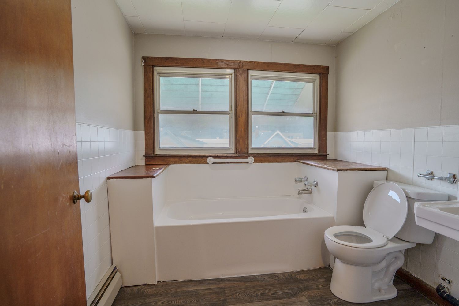 Bathroom with white soaking tub, toilet, tile wainscoting, two windows, and dark wood flooring.