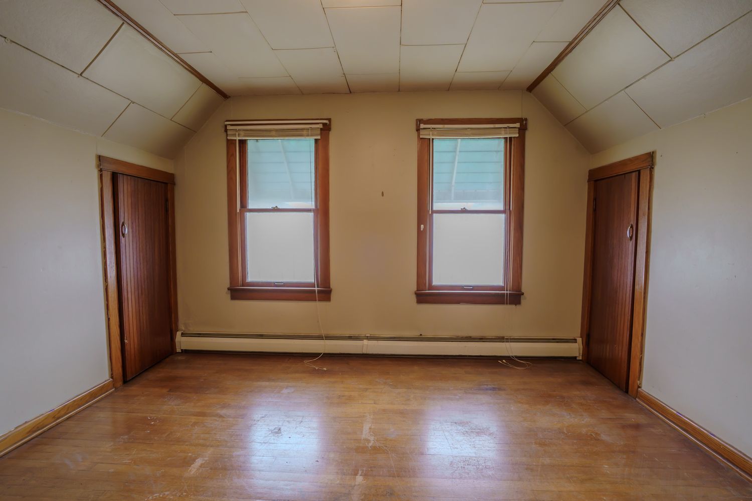 Empty attic room with hardwood floors, two windows, sloped ceiling, and two wooden doors.