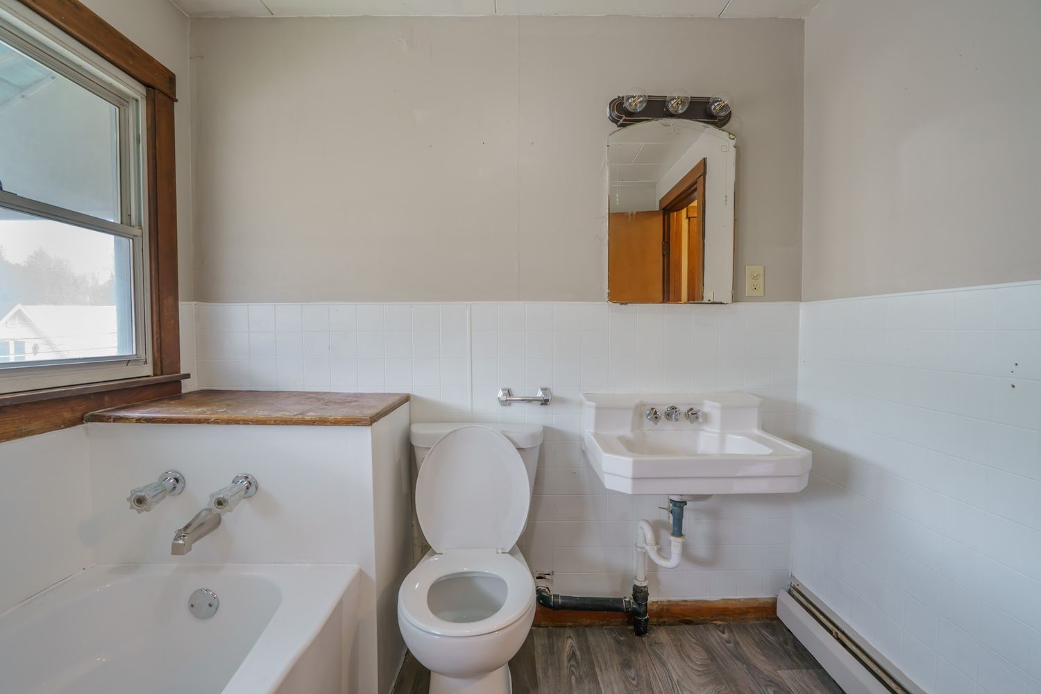 Vintage bathroom with white pedestal sink, toilet, soaking tub, wainscoting, wood floors, and mirror above sink.