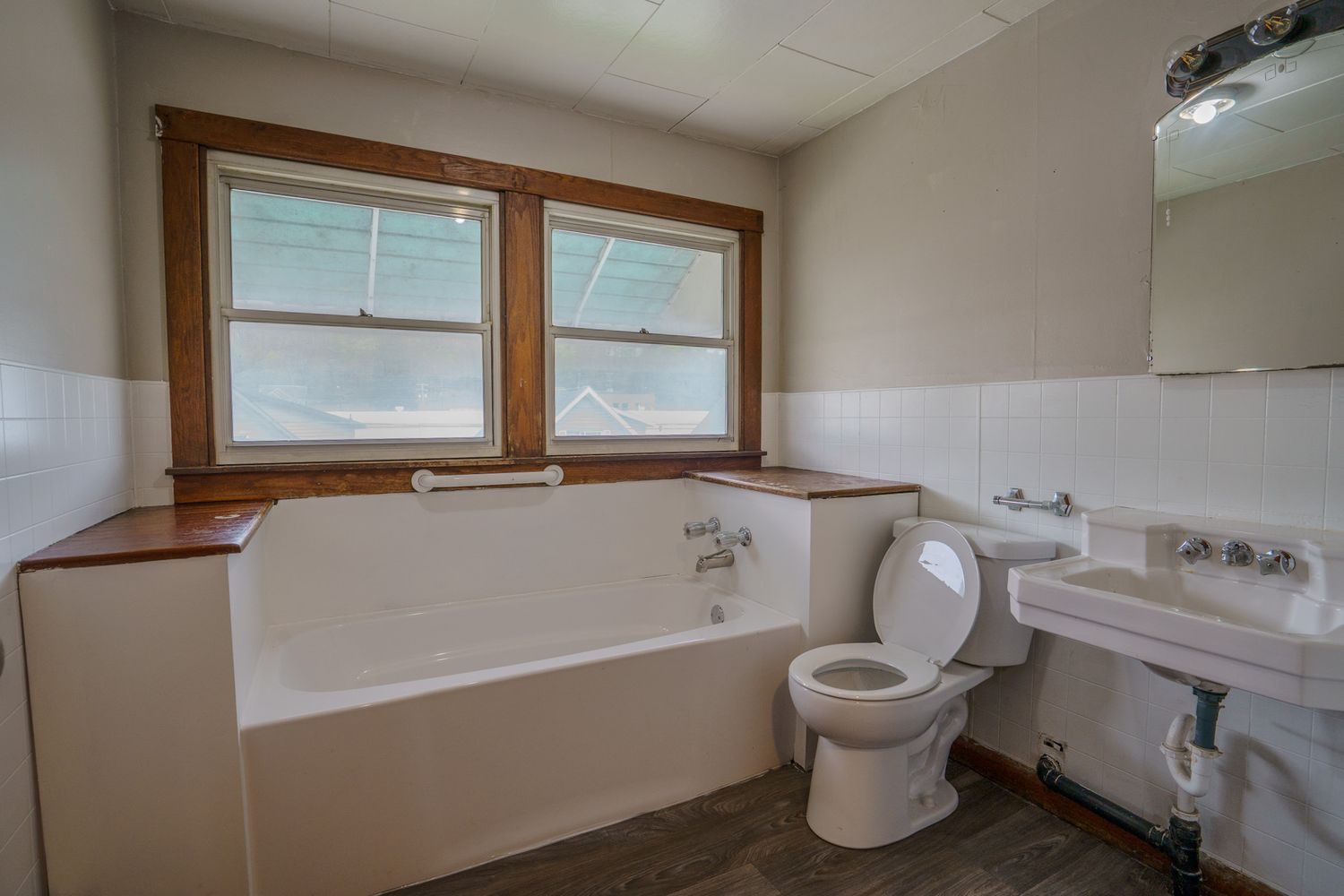 Vintage bathroom with white soaking tub, pedestal sink, toilet, and tiled walls with two wood-framed windows.