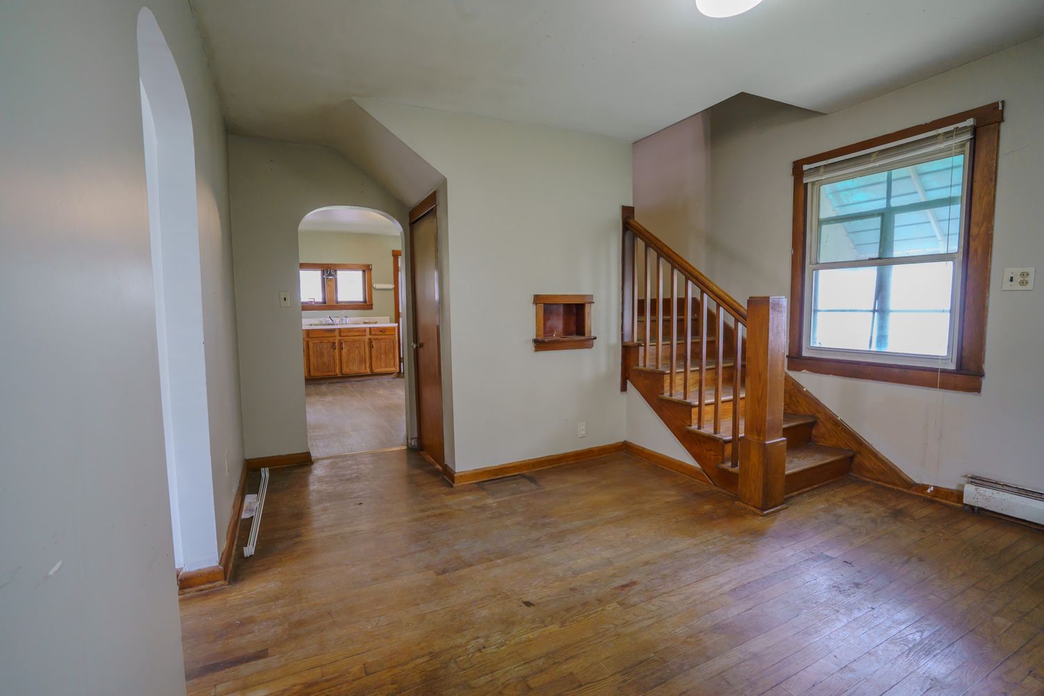Bright interior entryway with hardwood floors, wooden staircase, and arched hallway leading to kitchen.