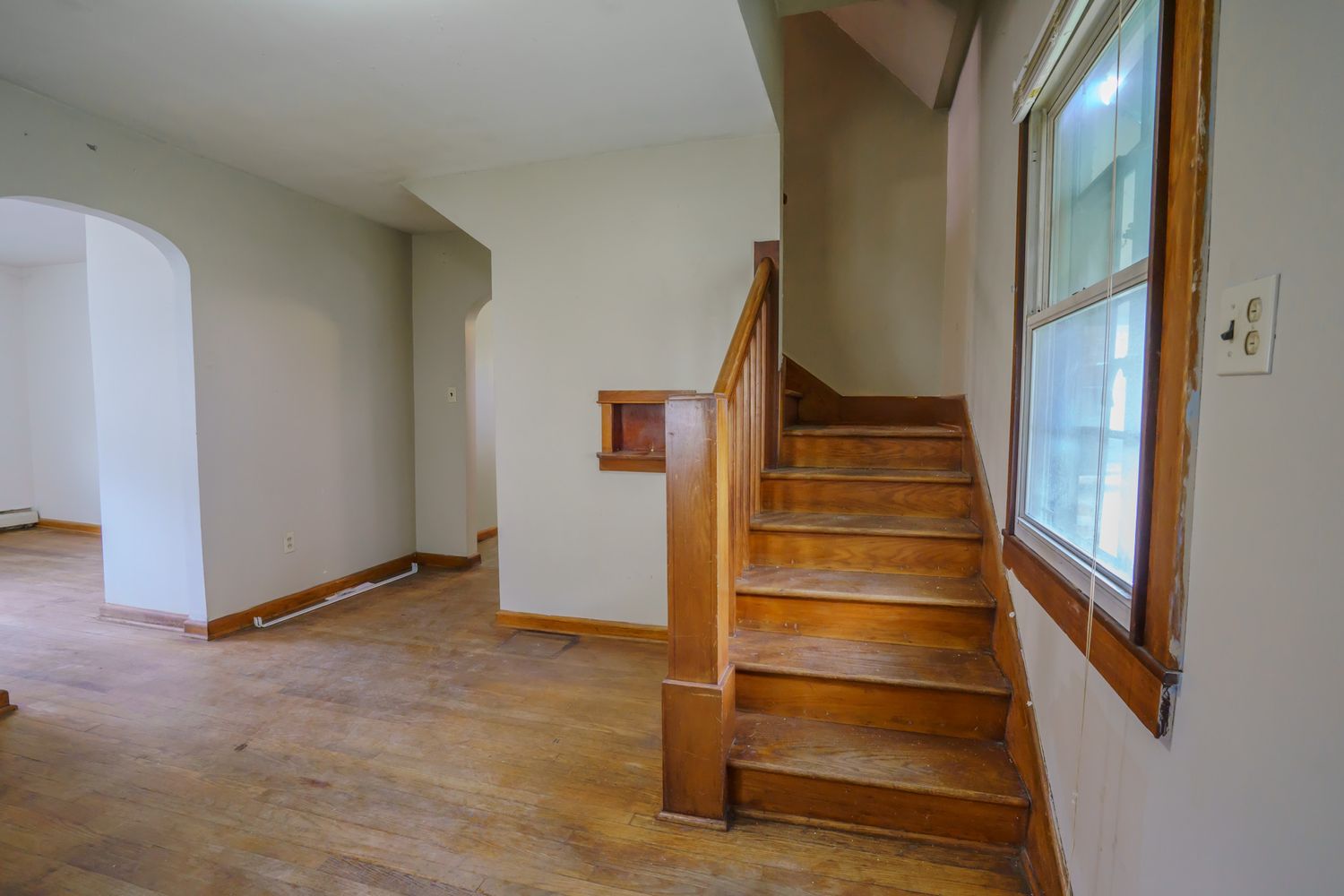Wooden staircase with handrail leads to upper level beside arched doorway and hardwood floors in bright home interior.