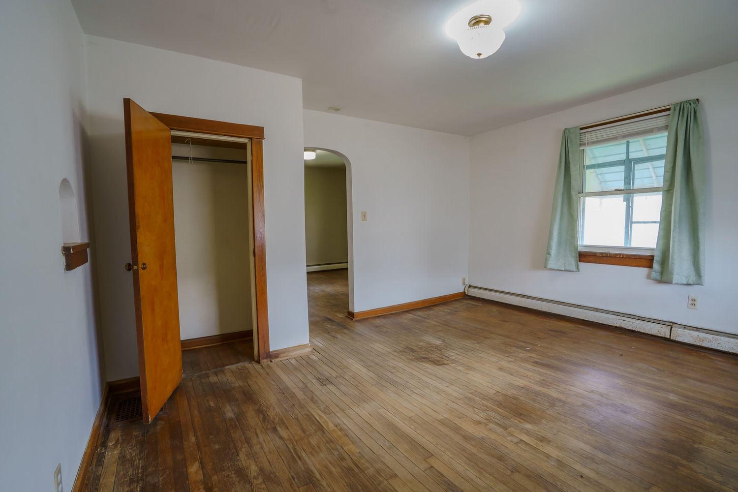 Empty bedroom with hardwood floors, open closet, baseboard heater, and a window with green curtains.