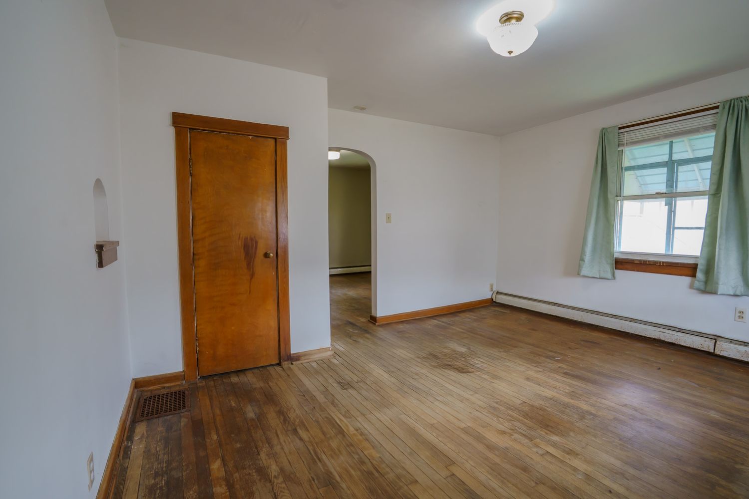 Empty bedroom with hardwood floors, baseboard heater, arched doorway, and green curtained window in a cozy home.