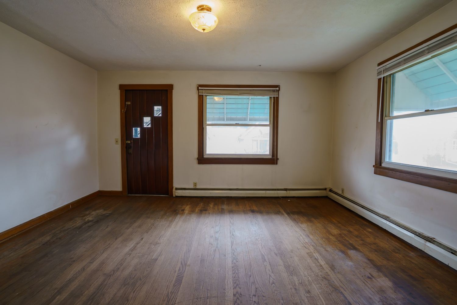 Empty room with hardwood floors, baseboard heater, two windows, and a dark wood interior door.