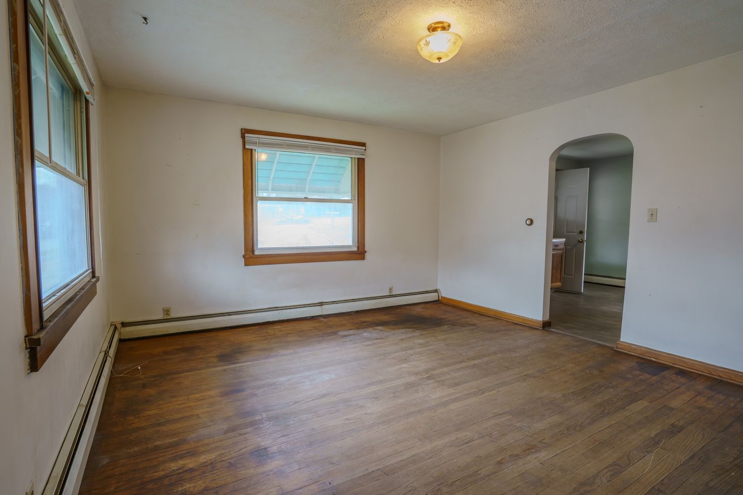 Empty room with hardwood floors, white walls, baseboard heater, arched doorway, and a window with natural light.