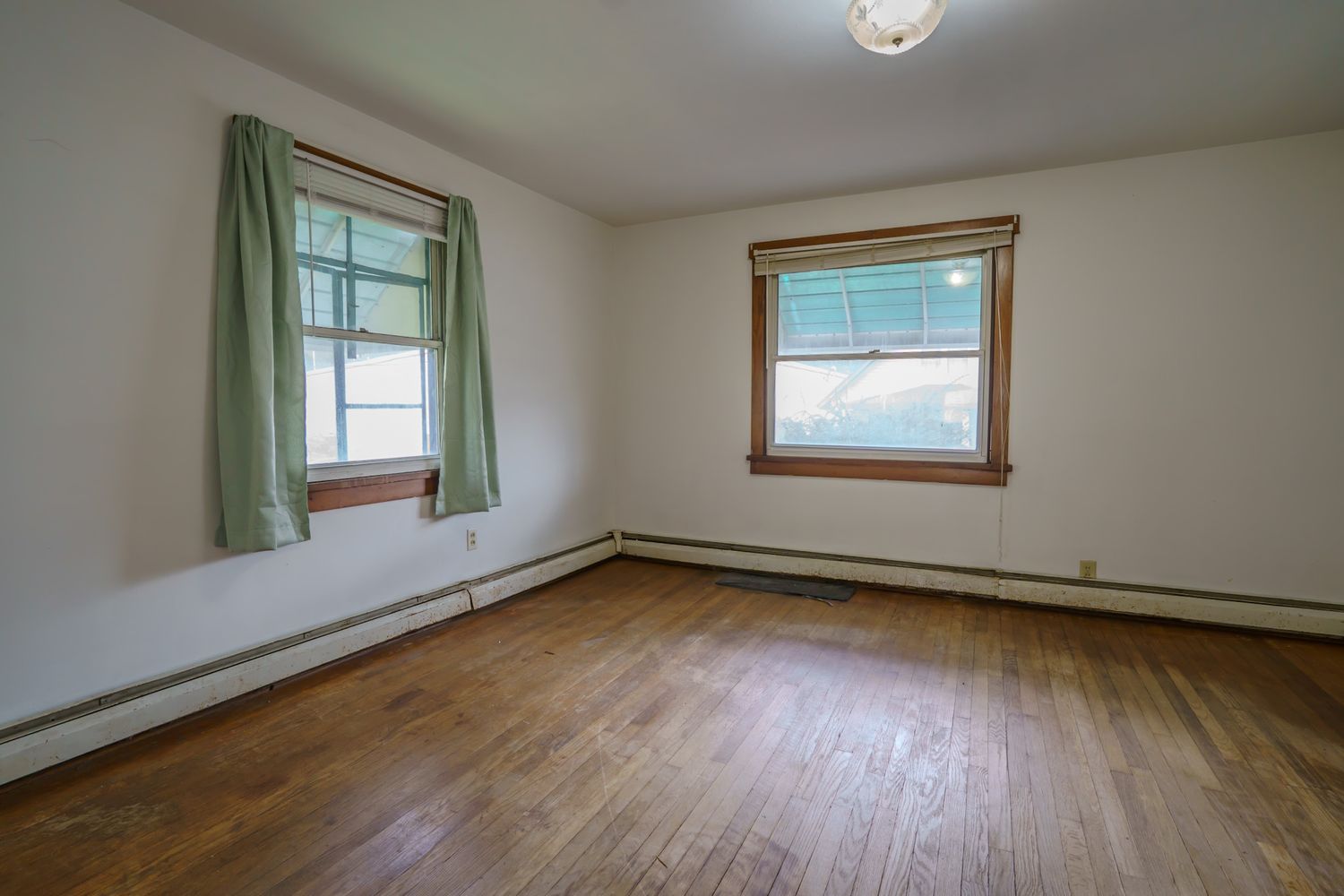 Empty room with hardwood floors, two windows with green curtains, and baseboard heating in a classic home.