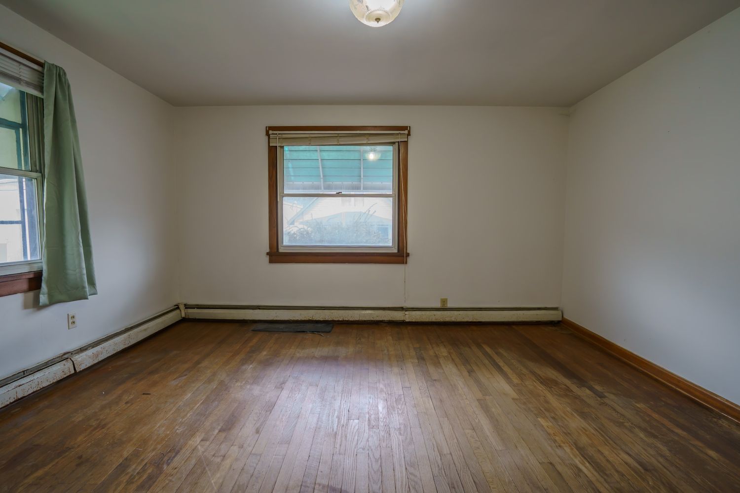Empty bedroom with hardwood floors, baseboard heater, and a single wood-framed window with green curtains.