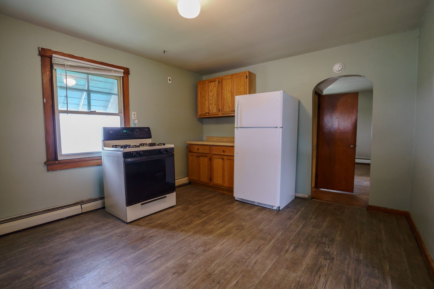 Sparse kitchen featuring gas range, white refrigerator, wood cabinets, and arched doorway with hardwood floors.