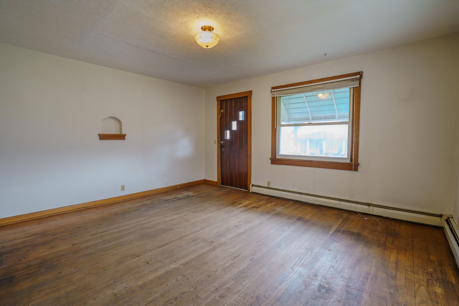 Empty room with hardwood floors, baseboard heater, wooden front door, and a small window letting in natural light.