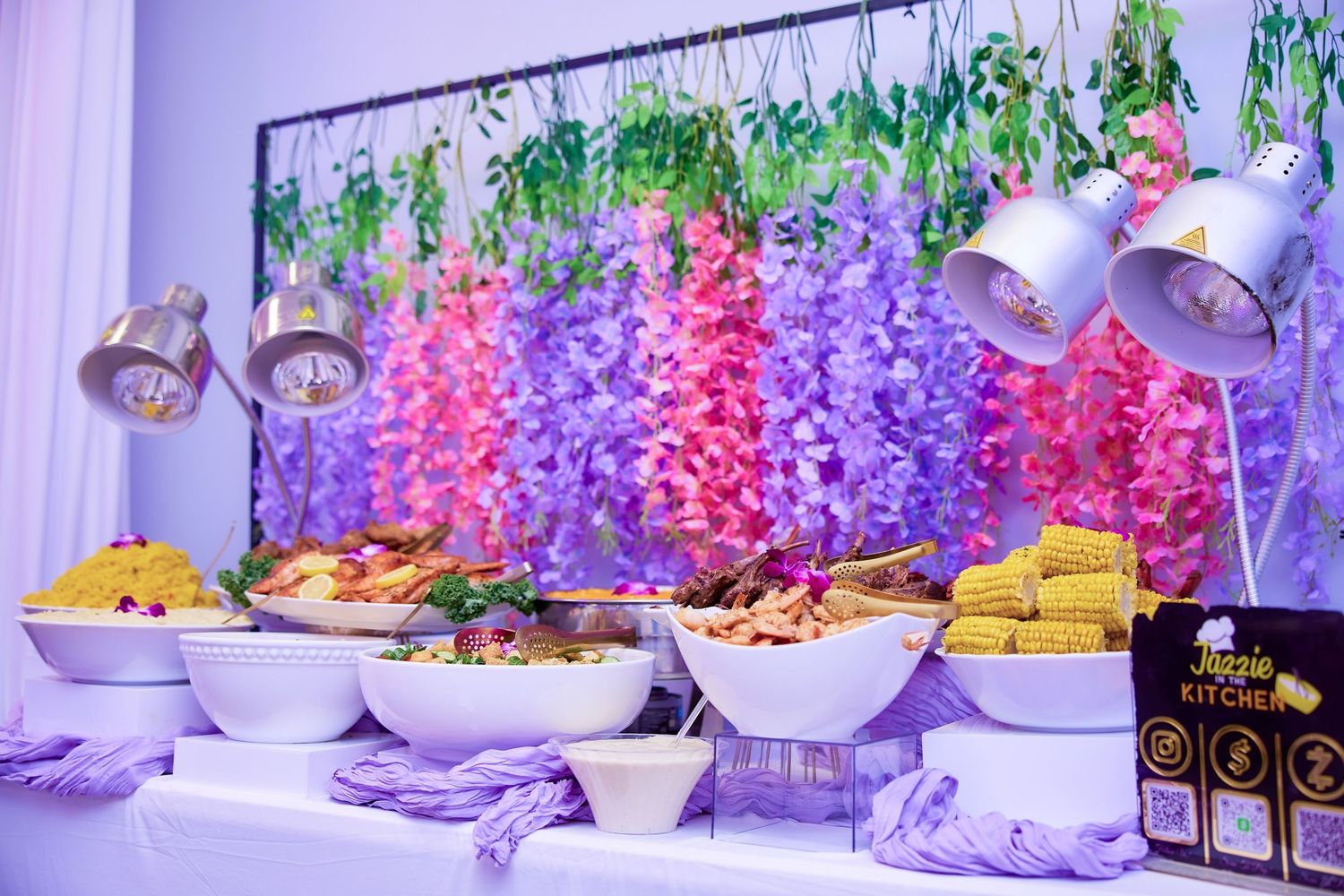 Elegant buffet spread with seafood and corn under heat lamps with floral backdrop.