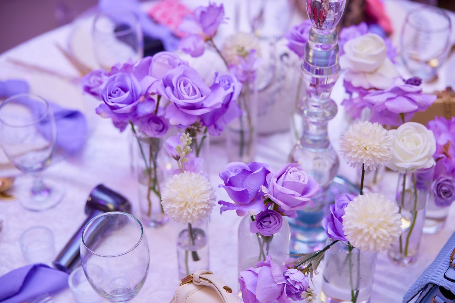 Purple and white floral centerpiece with roses and dahlias in glass vases on table.