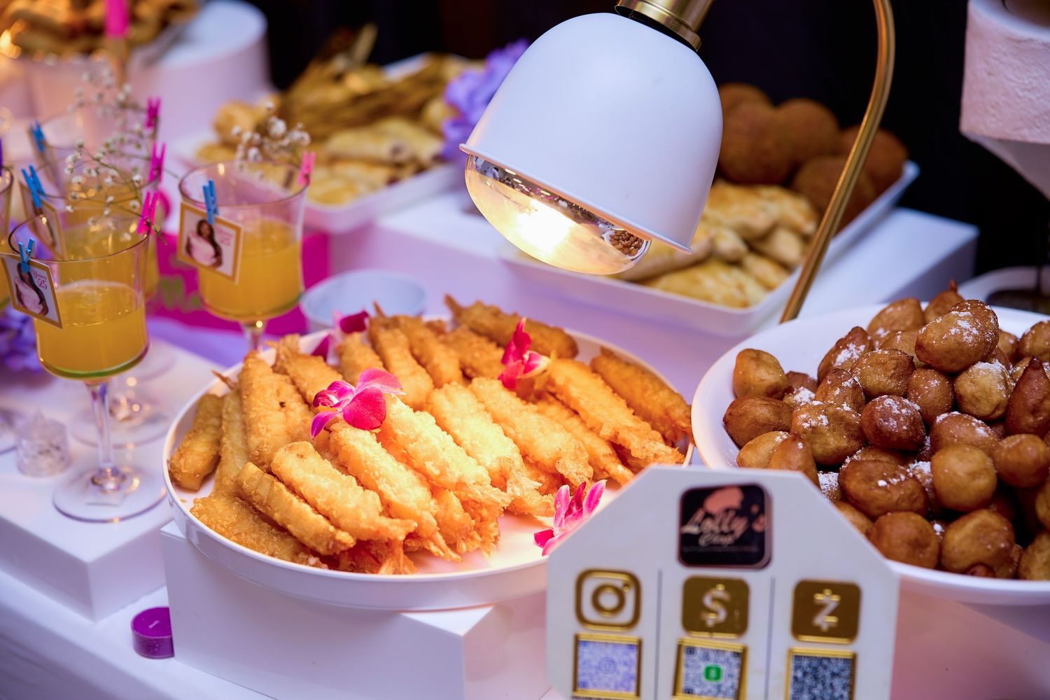 Elegant party food spread featuring fried appetizers, donut holes, and fresh juices displayed under warm lighting.