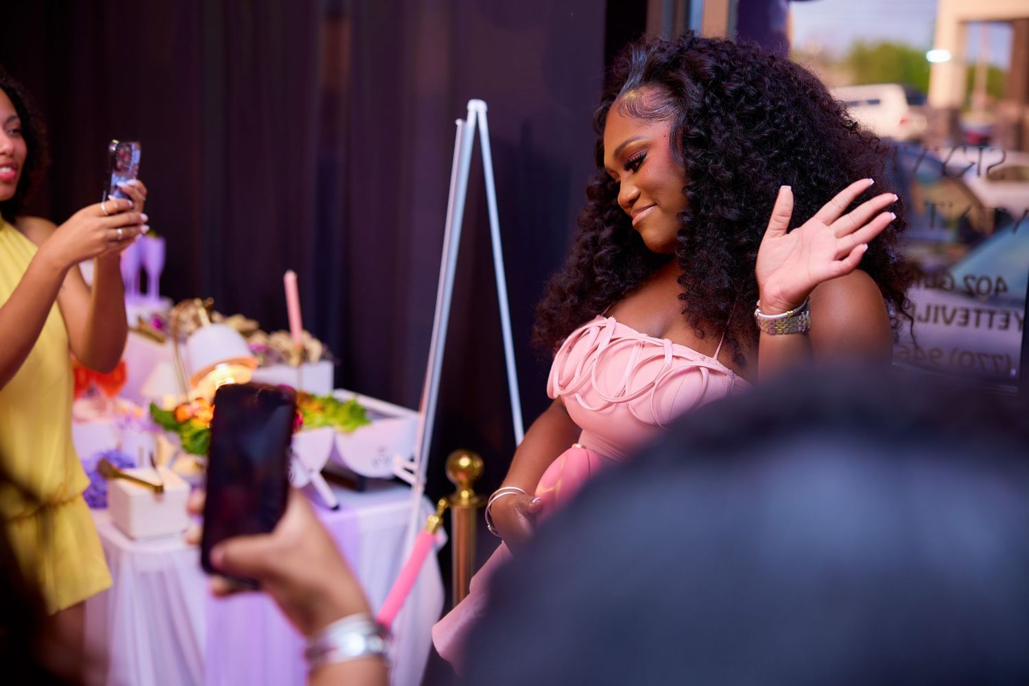 Woman in pink dress waving while guests photograph her at outdoor birthday celebration.
