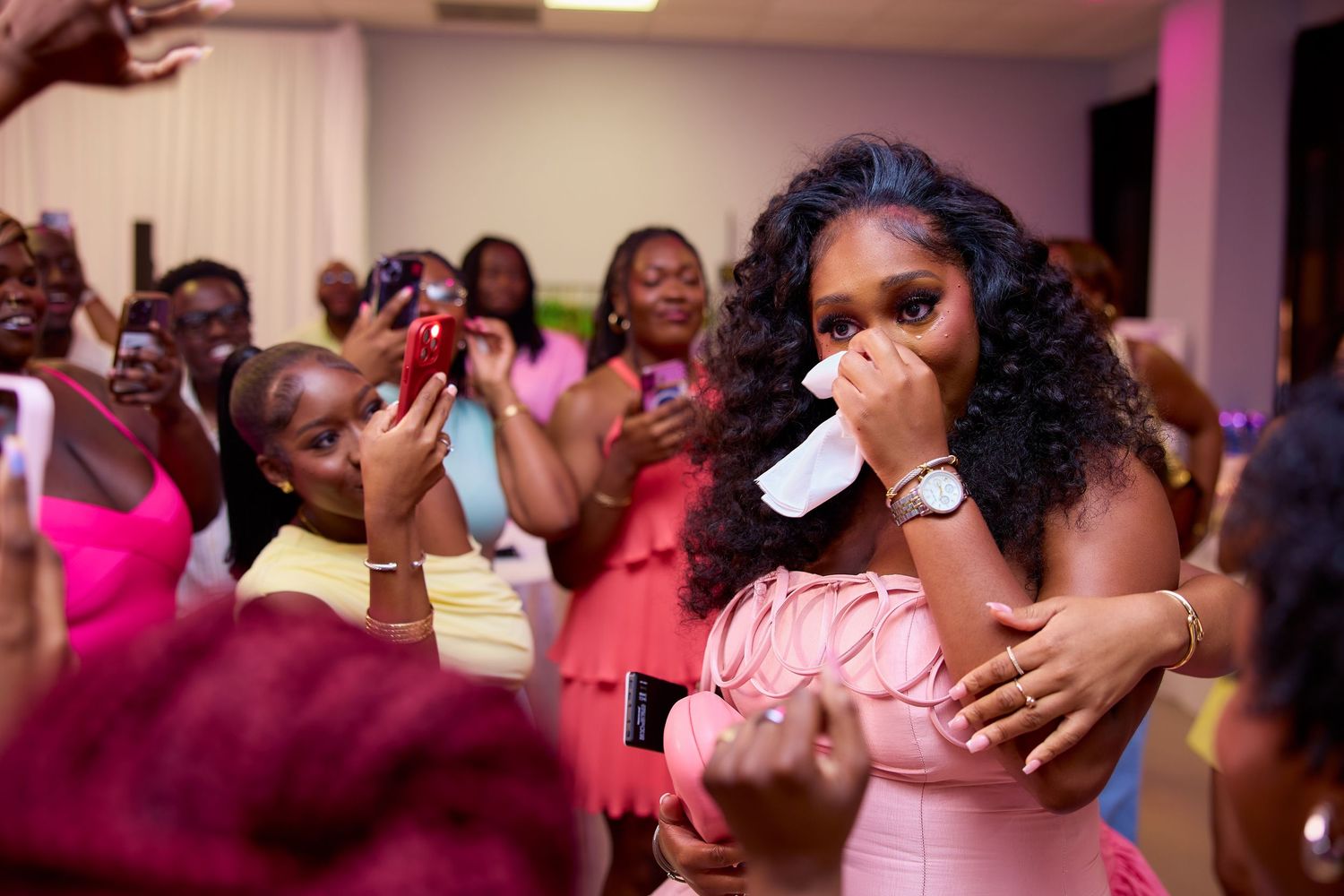 Emotional woman in pink dress wipes tears surrounded by celebrating guests at a surprise party.