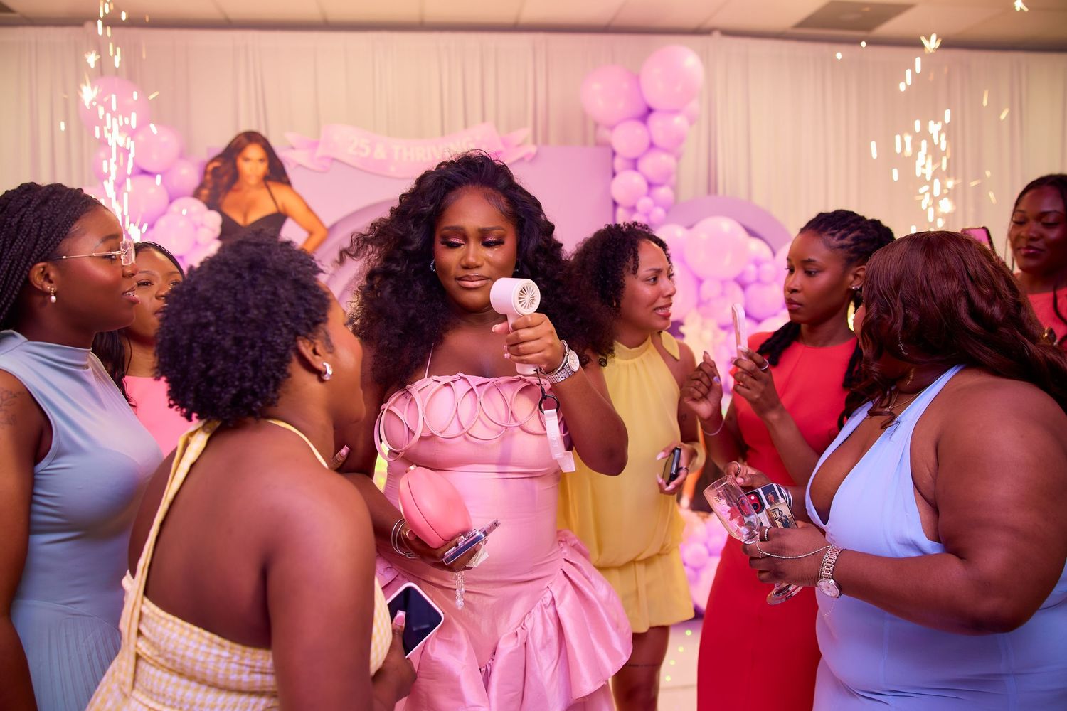 Birthday woman in pink dress holds microphone surrounded by guests at her party celebration.