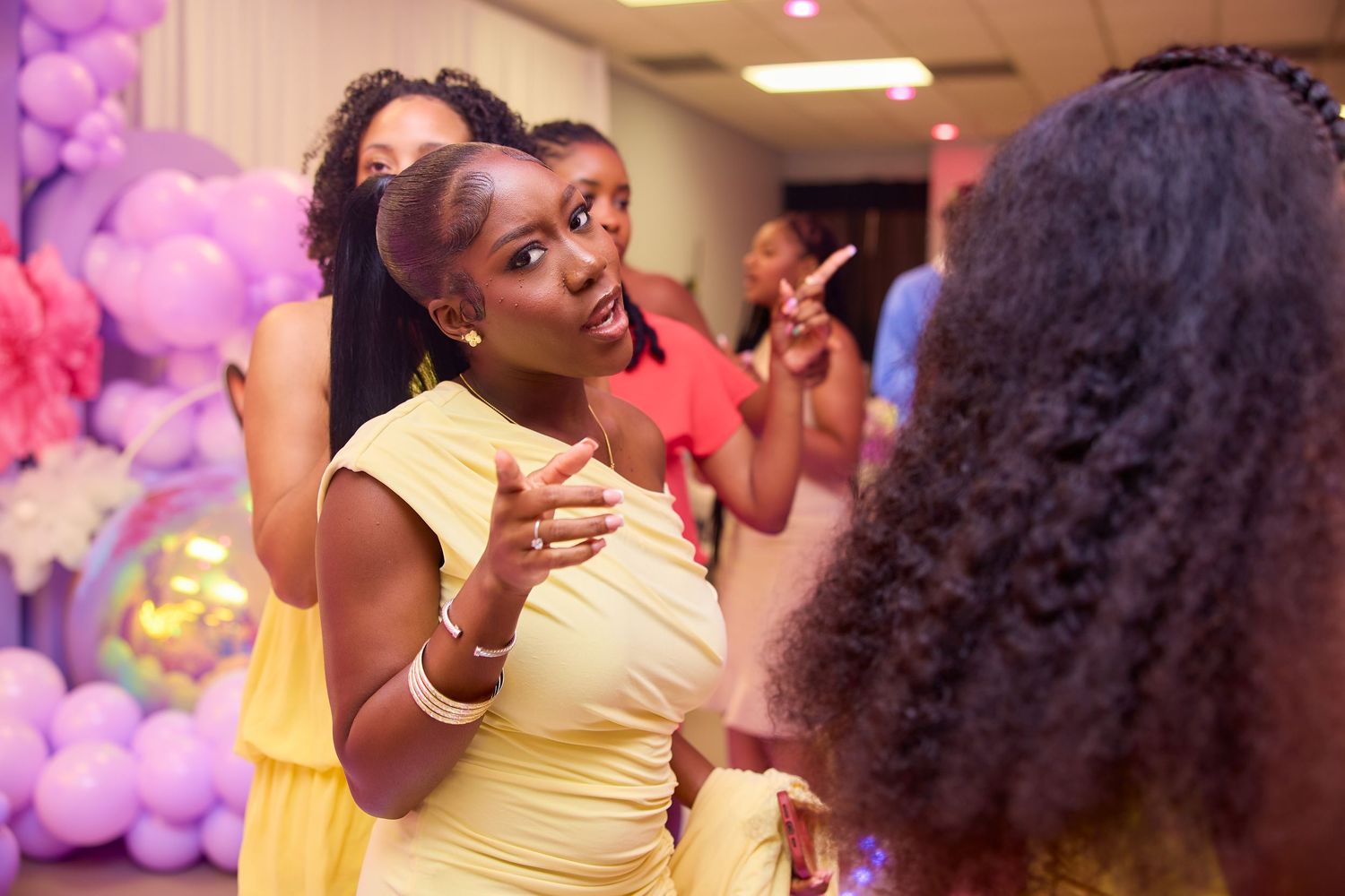 Women in yellow dress conversing animatedly near purple balloon decorations at party.