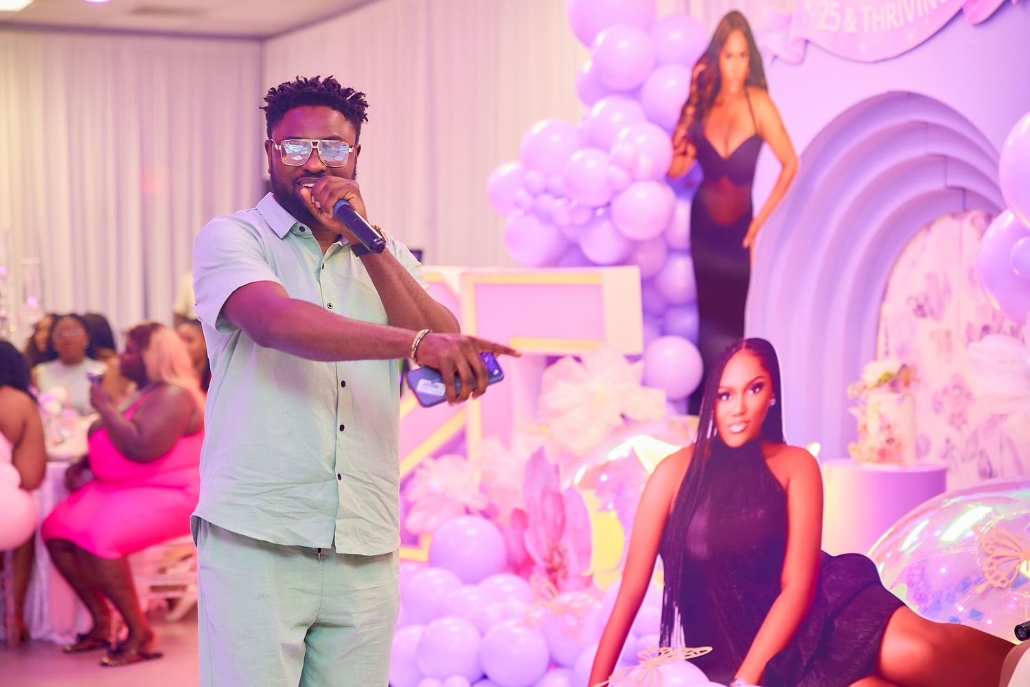Man speaking into microphone at purple balloon-decorated birthday party with cutout display.