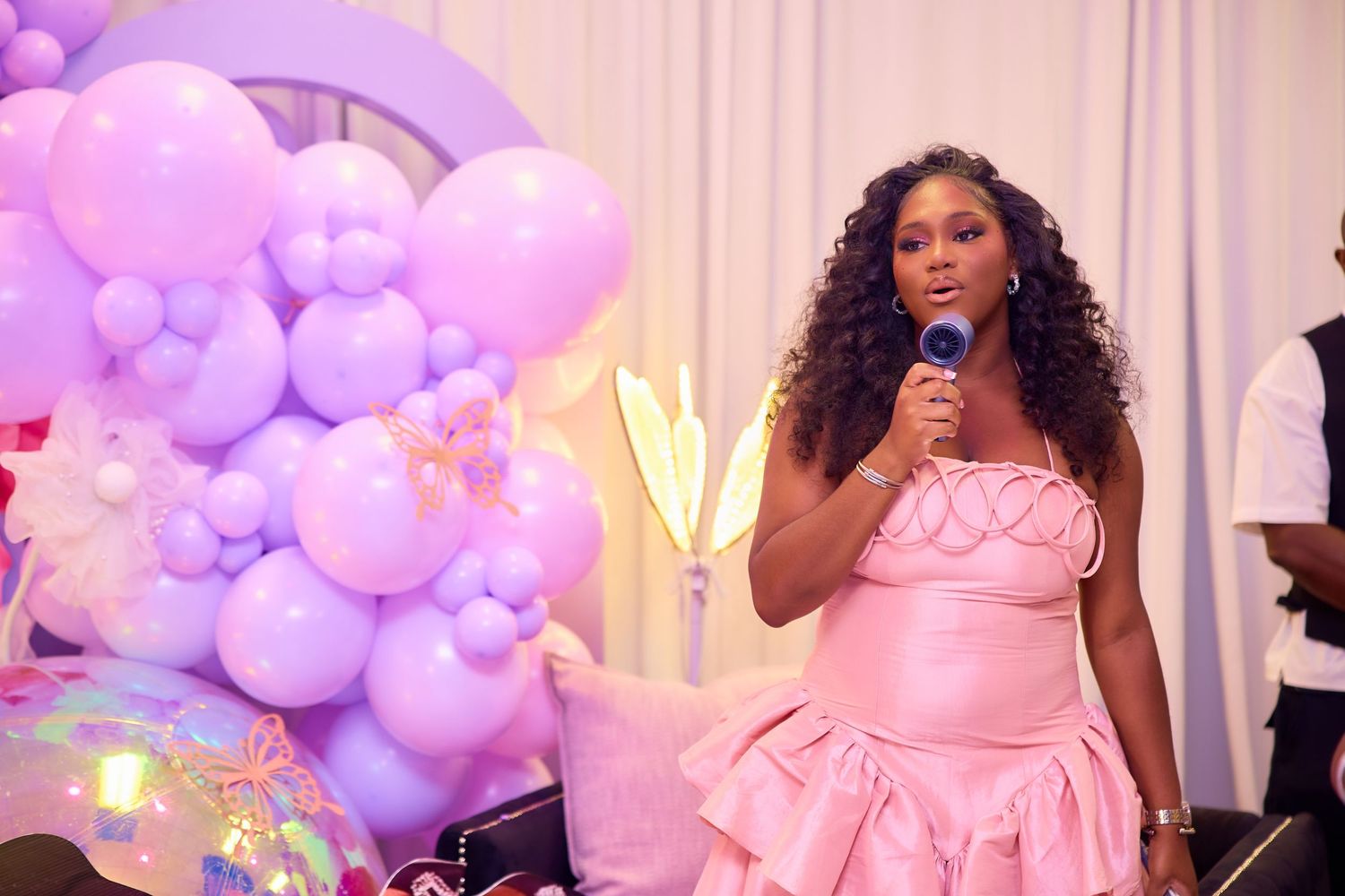 Woman in pink ruffled dress speaking into microphone at elegant party with purple balloon decorations.