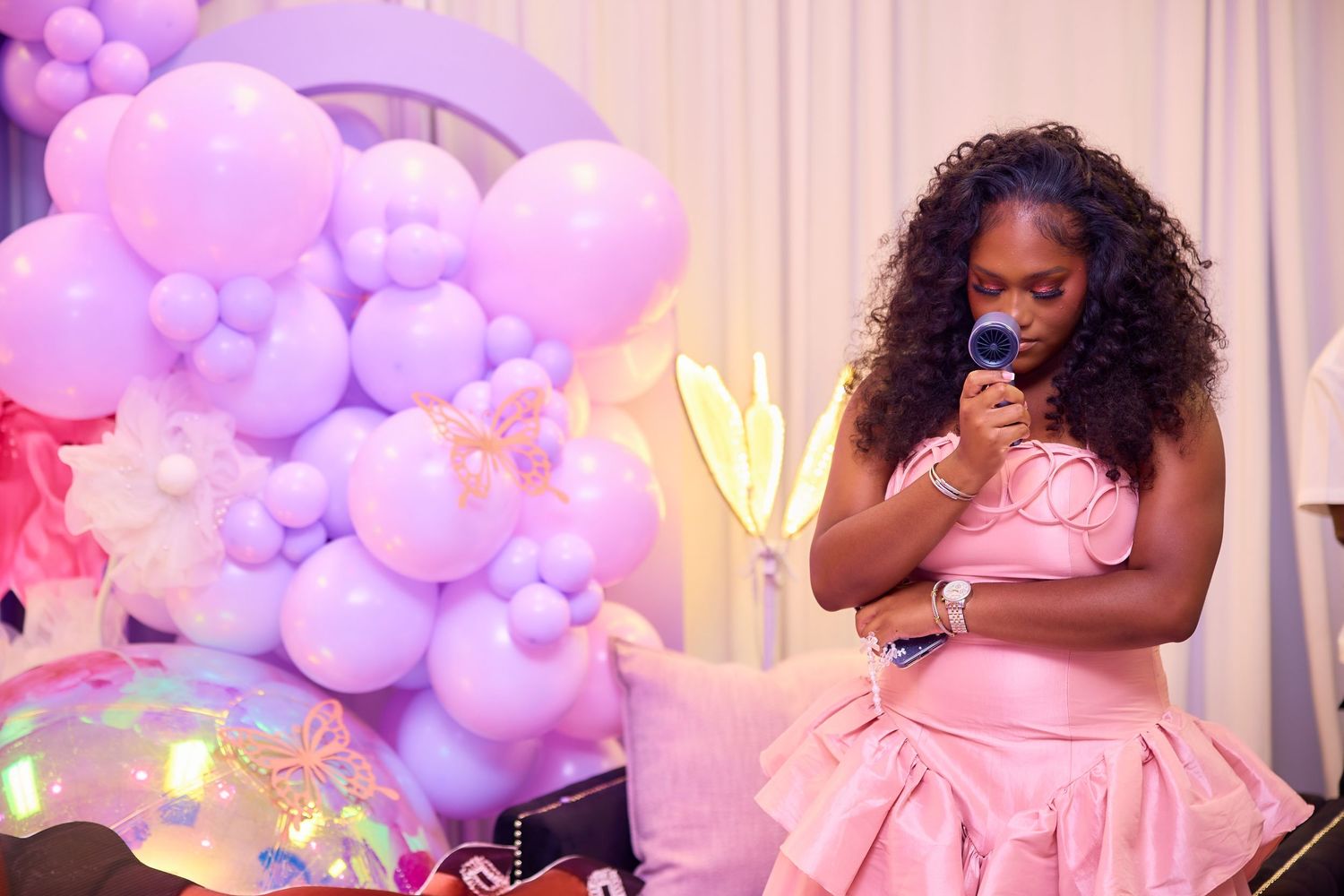 Young girl in pink ruffled dress speaks into microphone beside purple balloon arch at party.