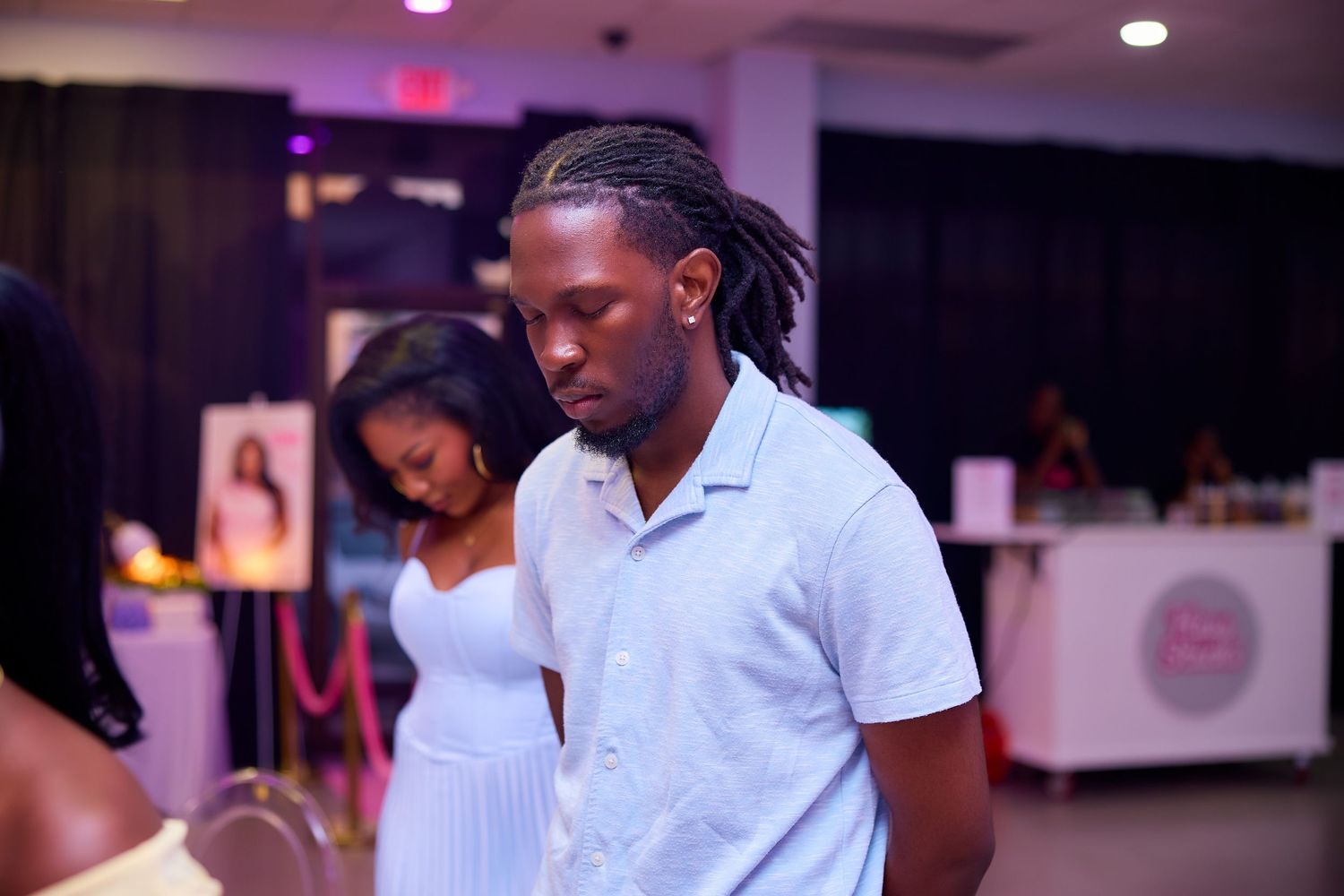 A young man with dreadlocks in a white shirt stands at an indoor celebration event.