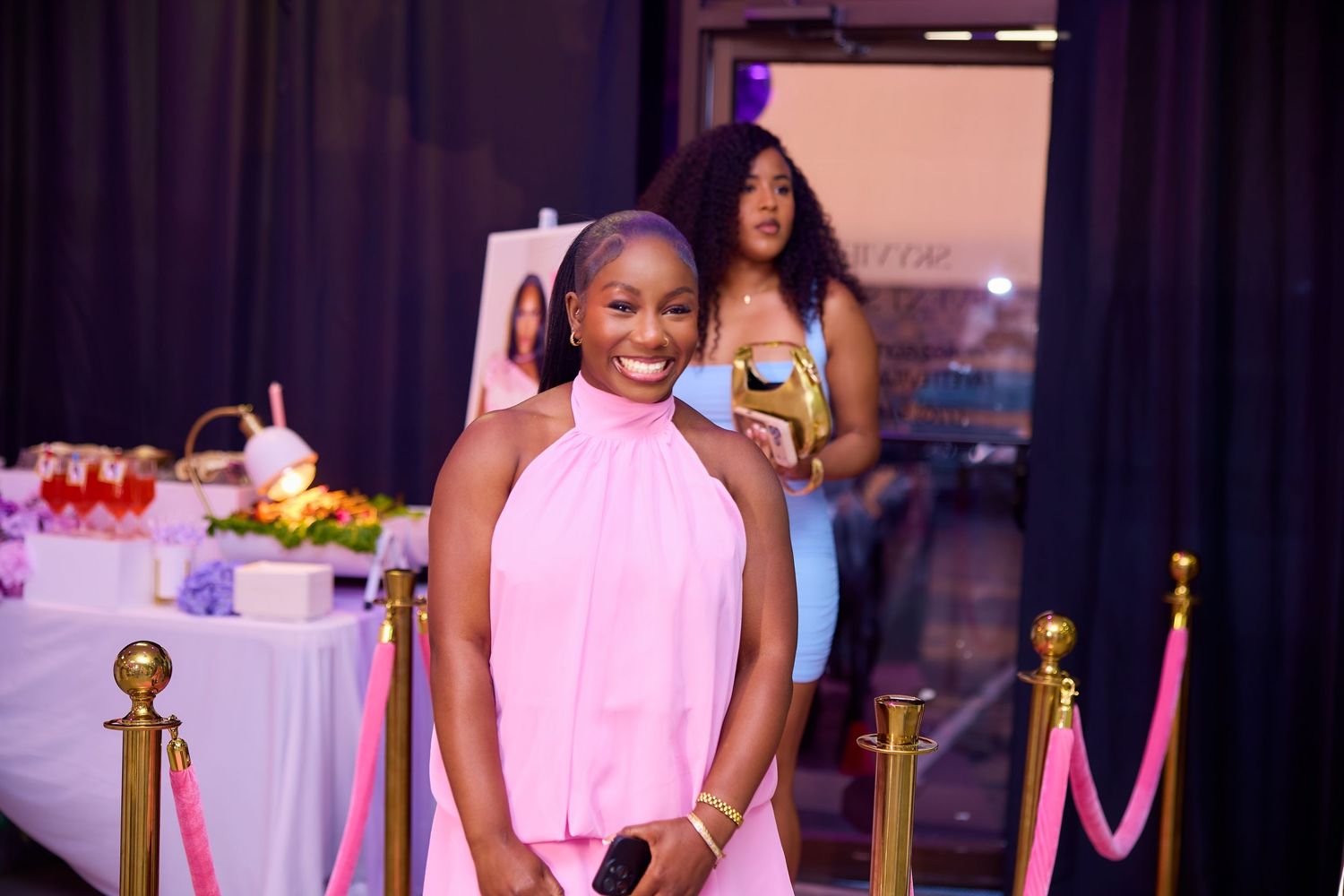 Smiling woman in pink halter dress stands on red carpet at formal event with gift table behind her.