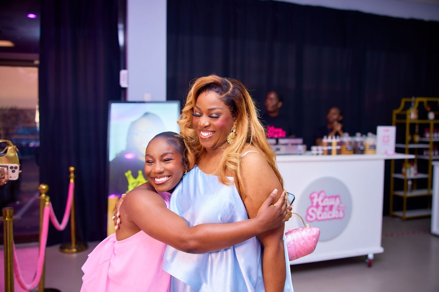 Two women in pastel dresses share a warm hug at an elegant indoor celebration event.