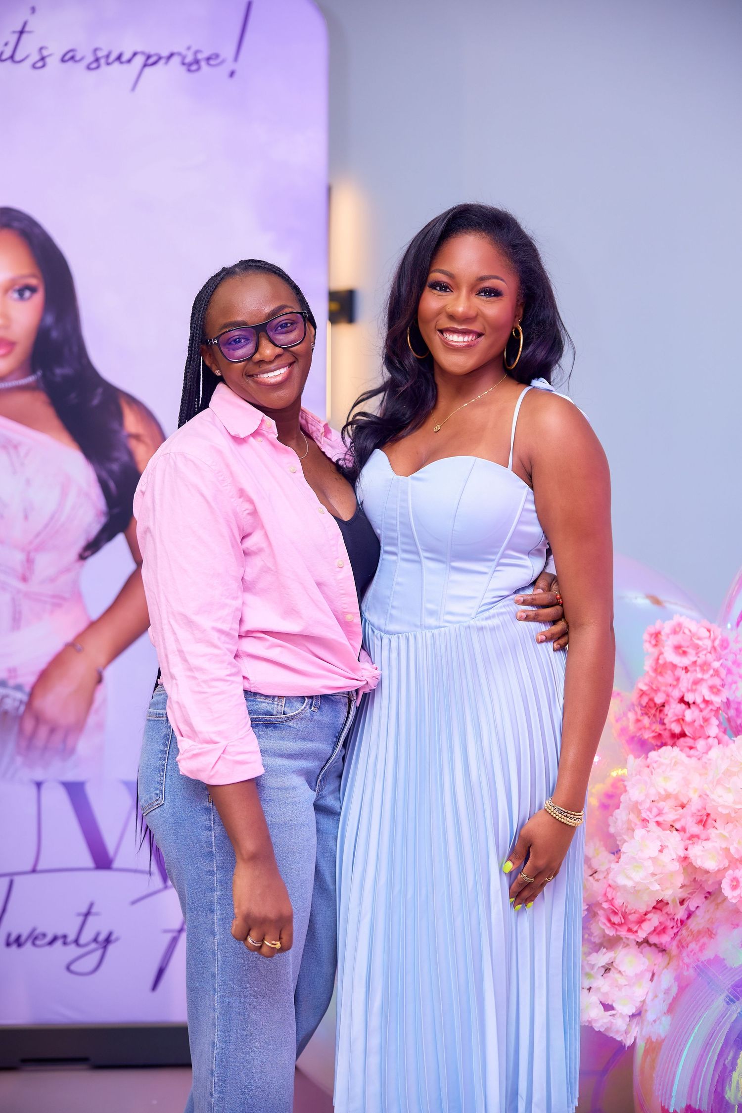 Two women pose together smiling at a surprise birthday party in front of a decorative banner.