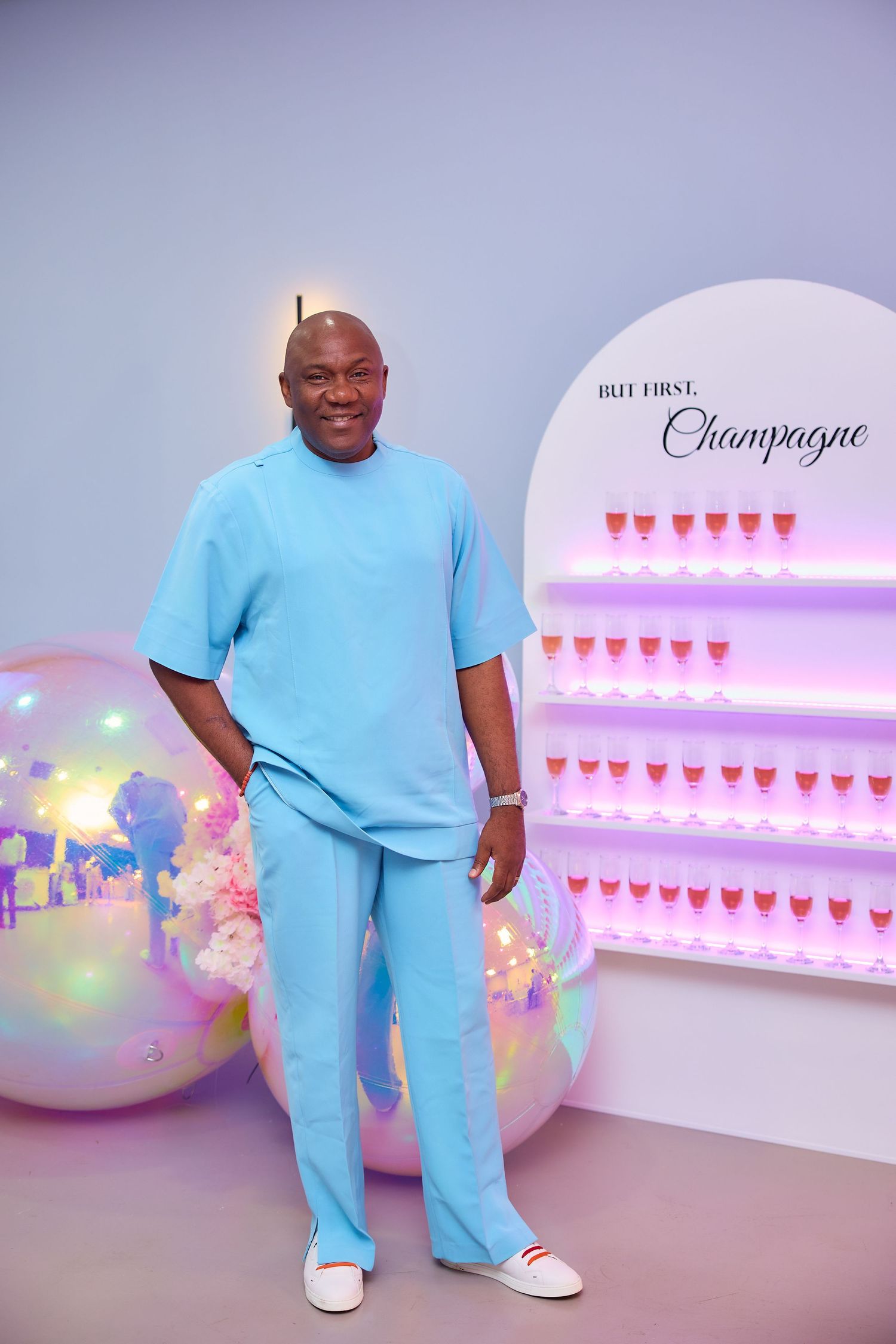 A smiling man in a turquoise outfit poses by a champagne wall display at a party.