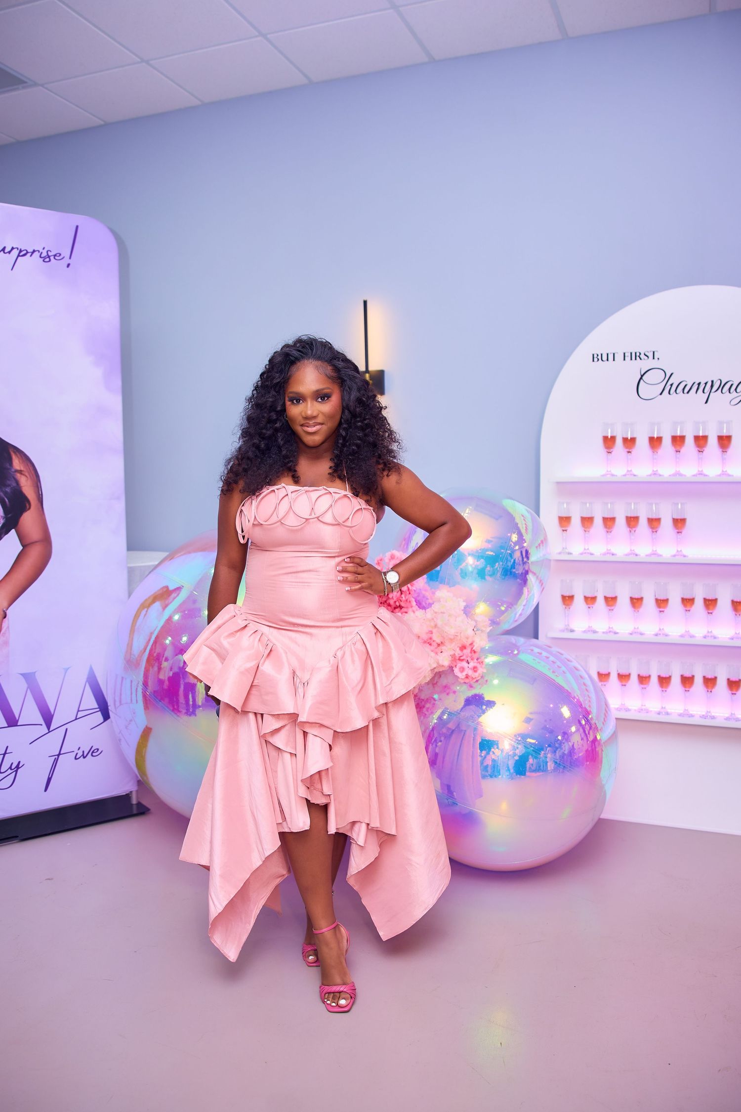 Elegant woman in pink ruffled gown poses at birthday party with champagne wall and balloon decor.