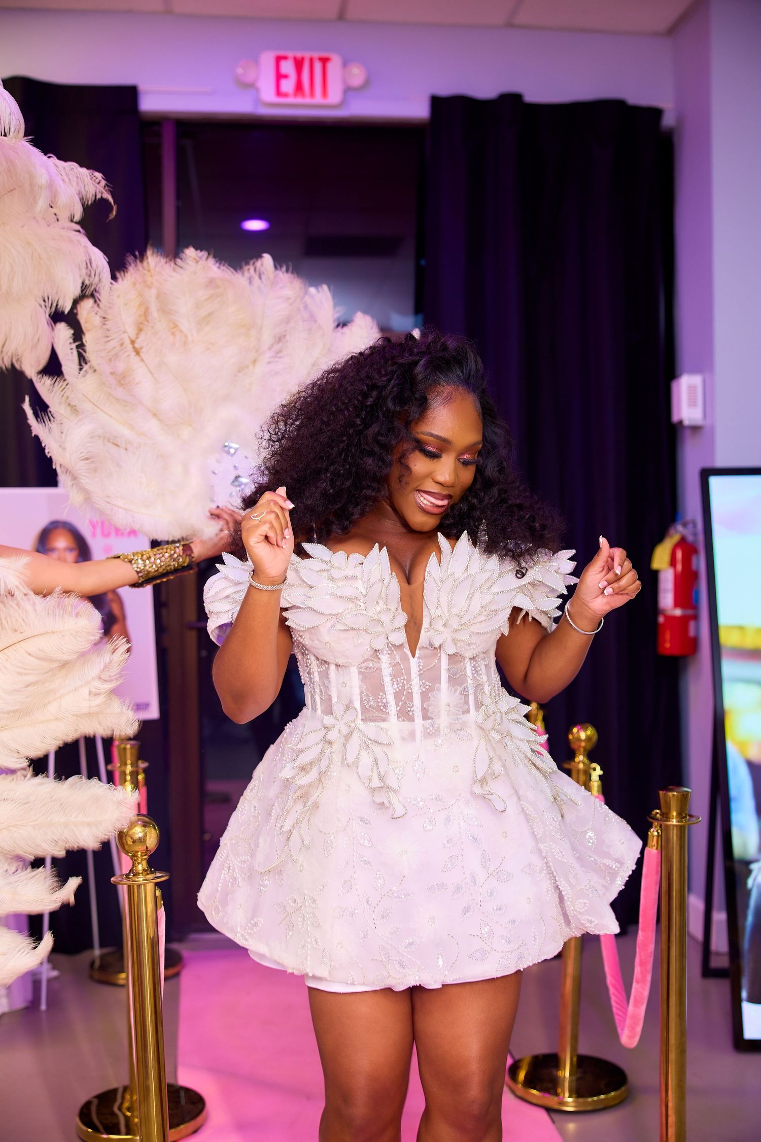 Woman in white feathered mini dress holding feather fan at elegant event with gold rope barriers.