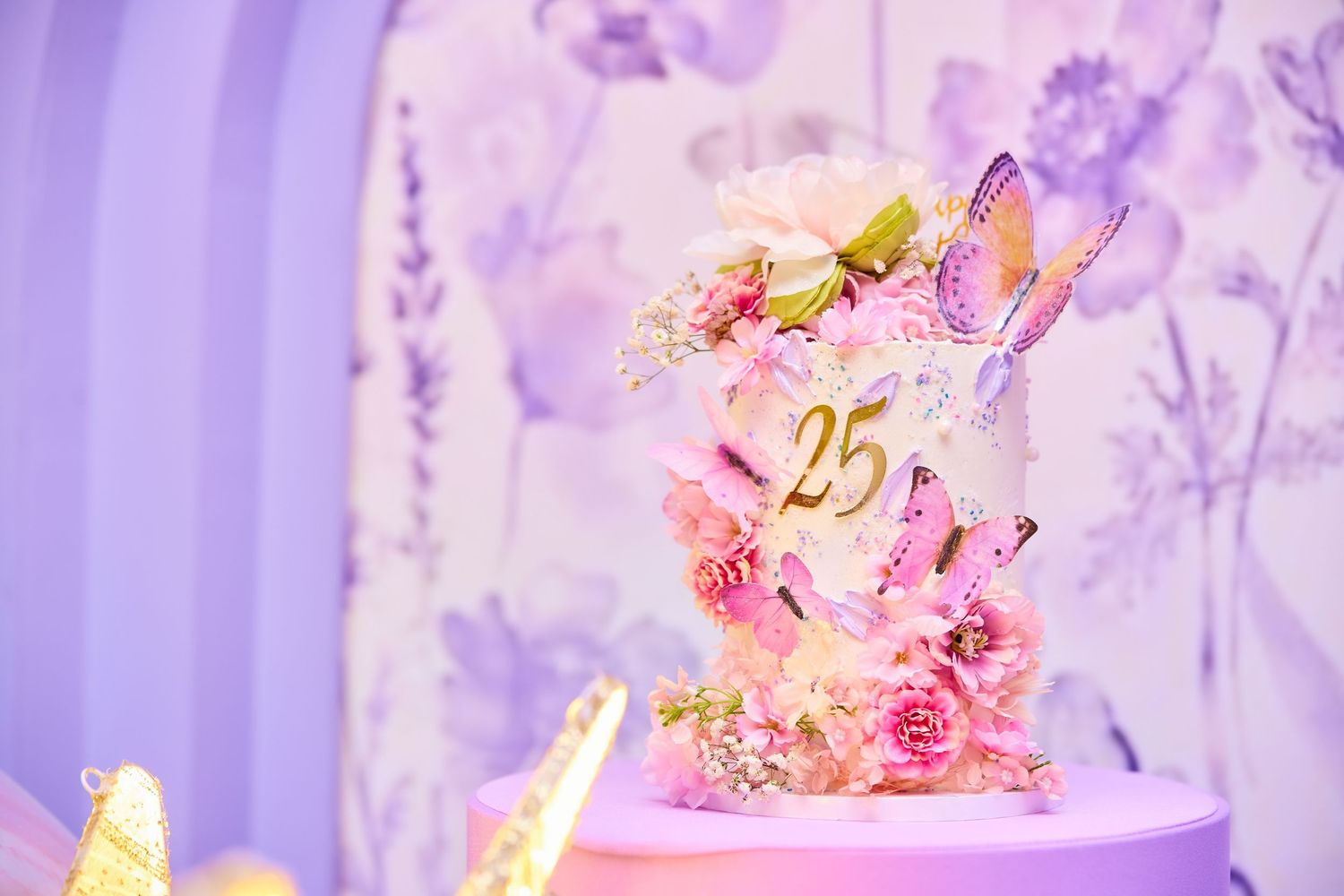 Pink butterfly-adorned 25th anniversary cake with floral decorations on a purple pedestal against a floral backdrop.