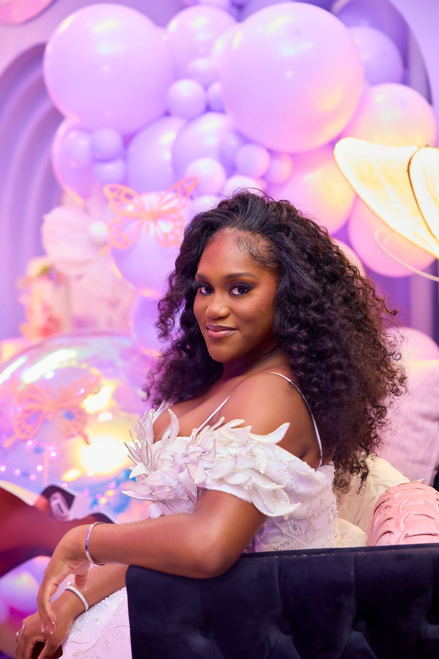Woman in white feathered dress smiles at purple balloon-decorated birthday celebration.