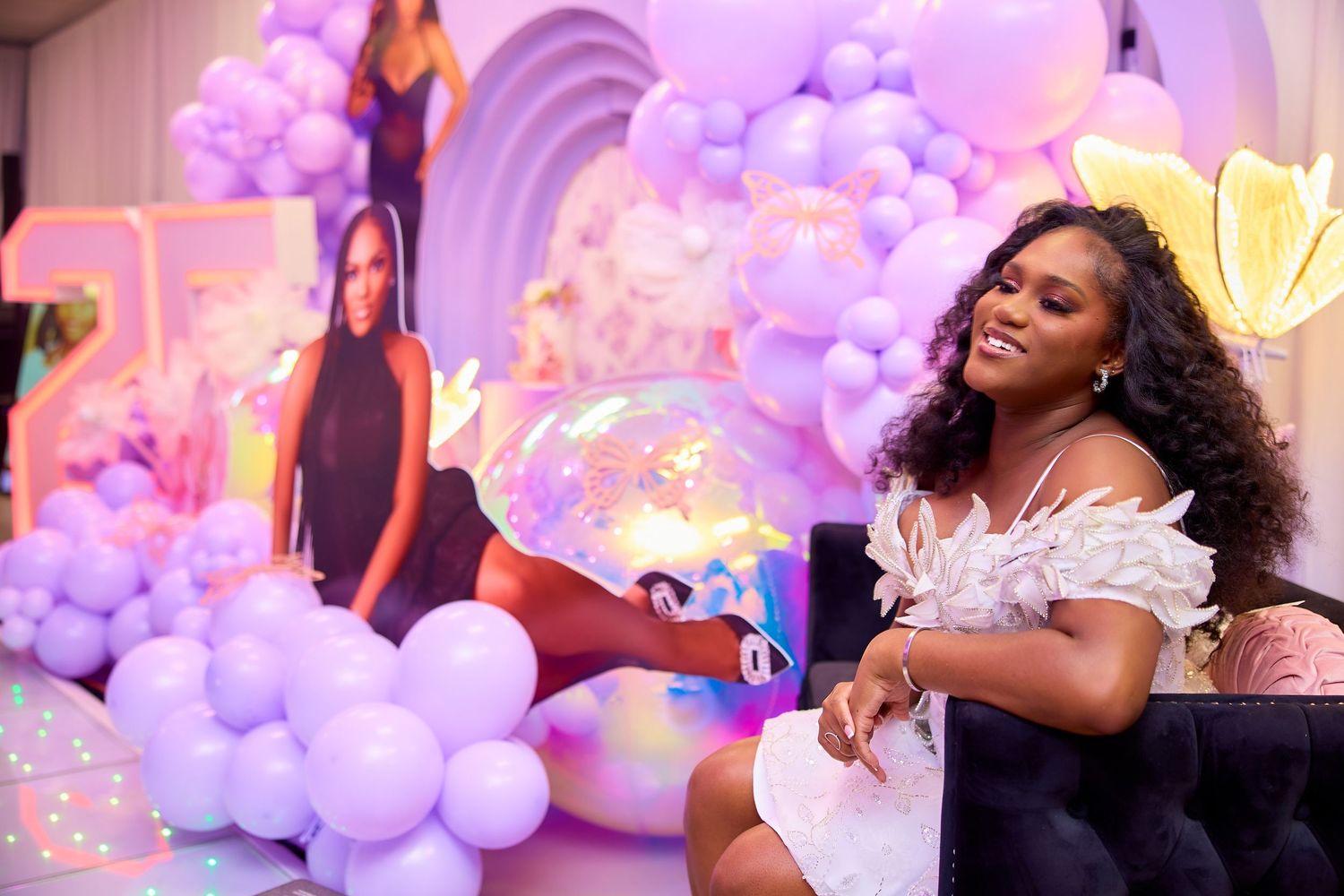 Birthday girl in white dress smiling beside purple balloon decorations and custom photo backdrop.