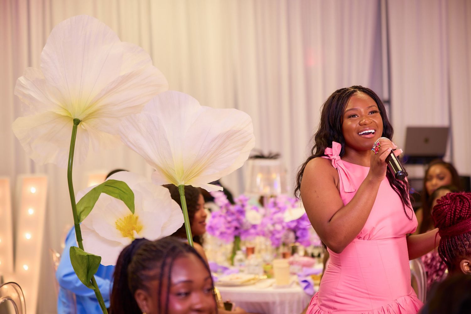 Woman in pink dress speaking into microphone at an elegant floral decorated event.
