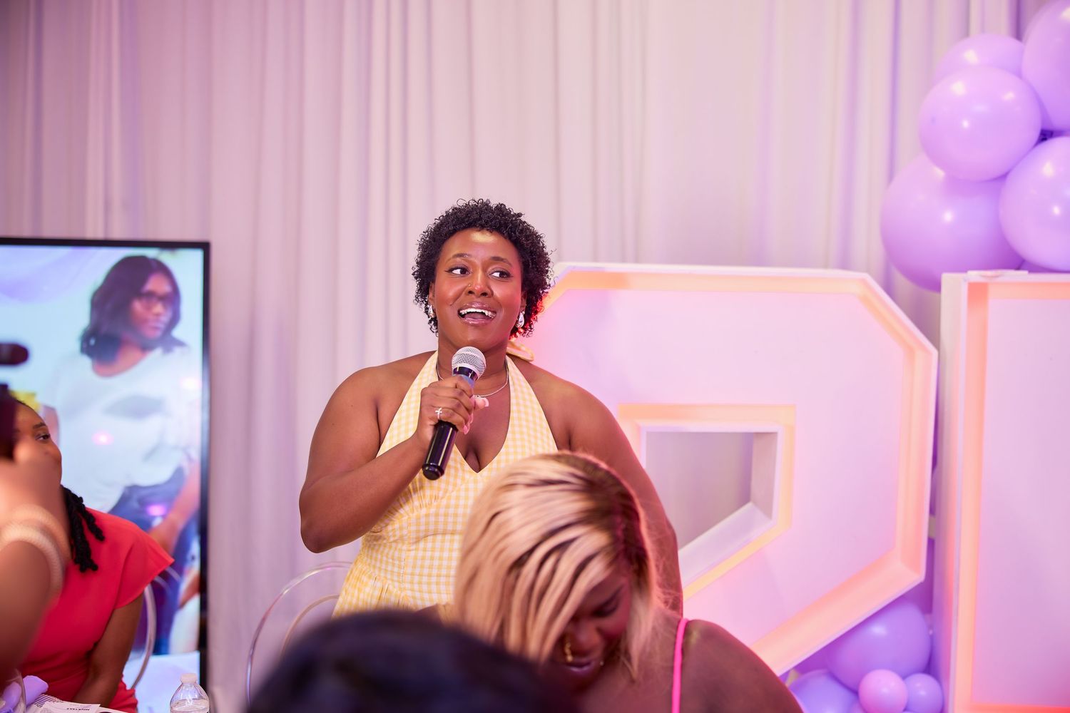 Woman speaking into microphone at a purple balloon decorated birthday celebration.