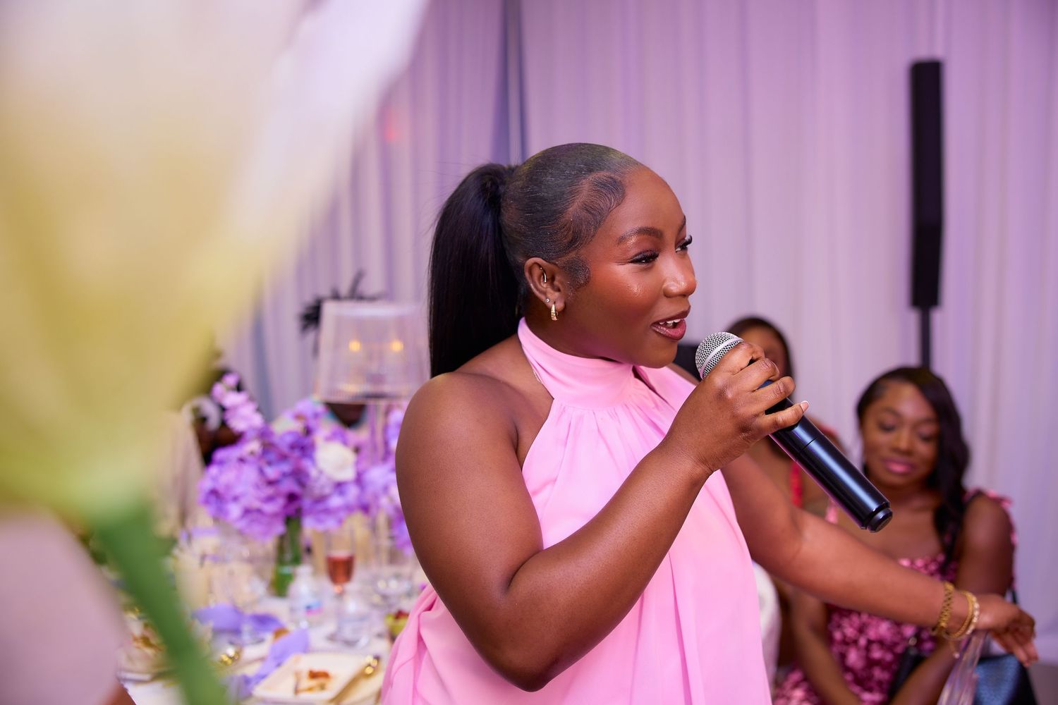 Woman in pink halter dress speaking into microphone at elegant purple-themed celebration event.