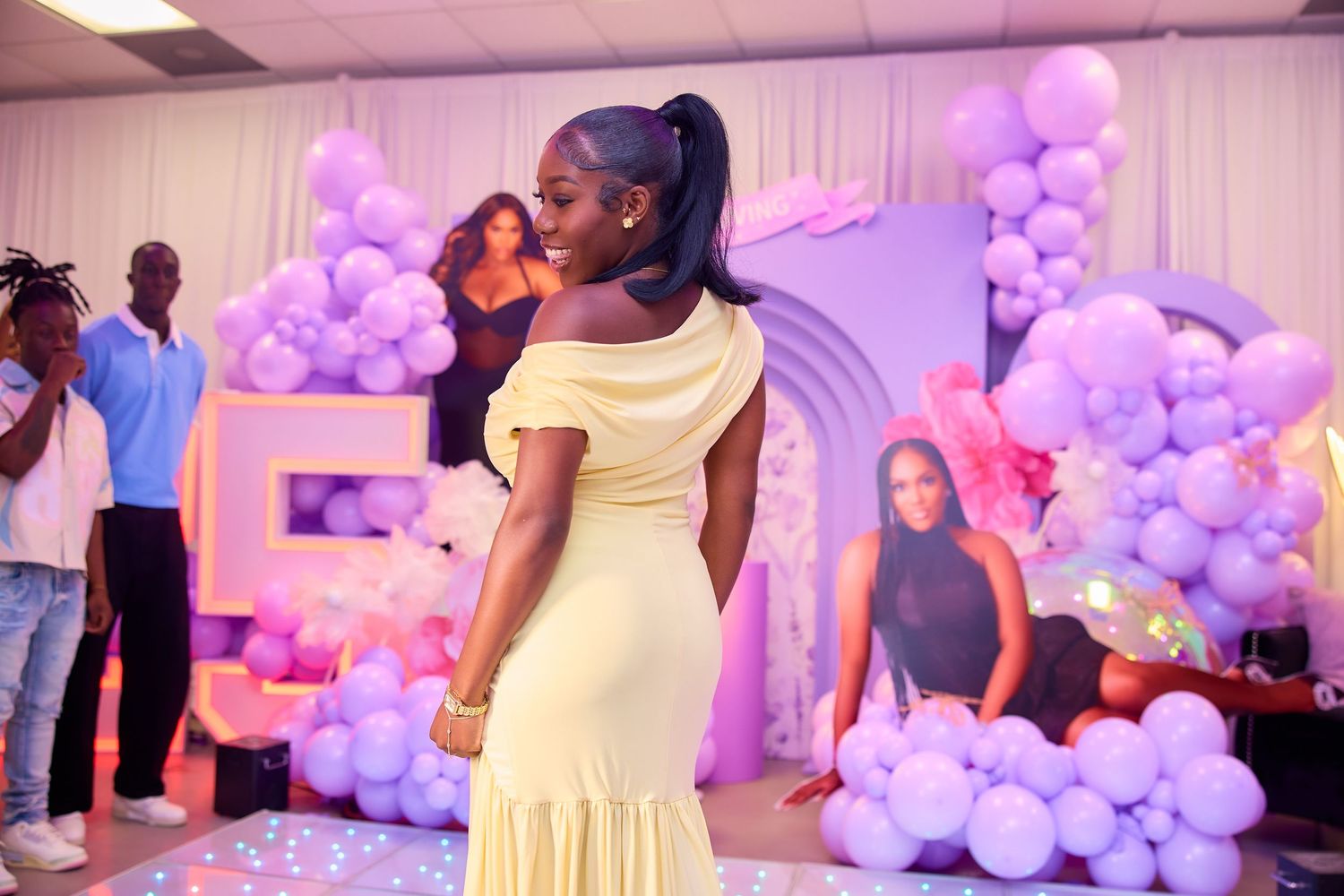 Woman in yellow dress poses in front of purple balloon decor and birthday backdrop.