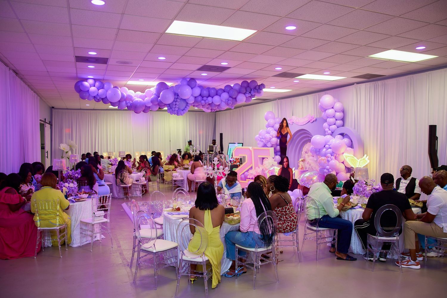 Elegant purple-themed event venue filled with guests seated at decorated tables under balloon arrangements.