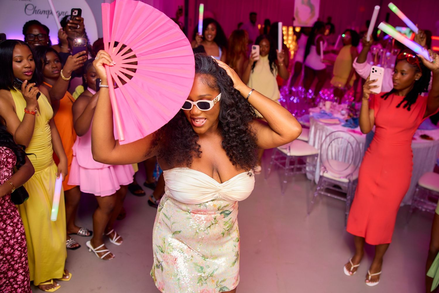 Woman in floral dress and sunglasses dancing with pink fan at vibrant birthday party.