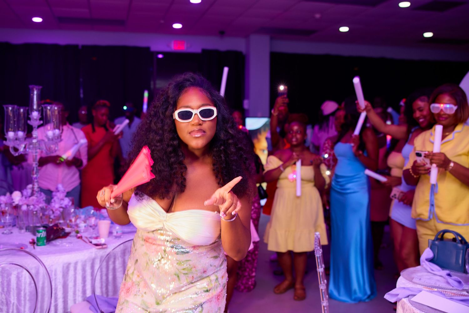 Woman in white sunglasses and floral outfit dancing at a vibrant purple-lit party event.