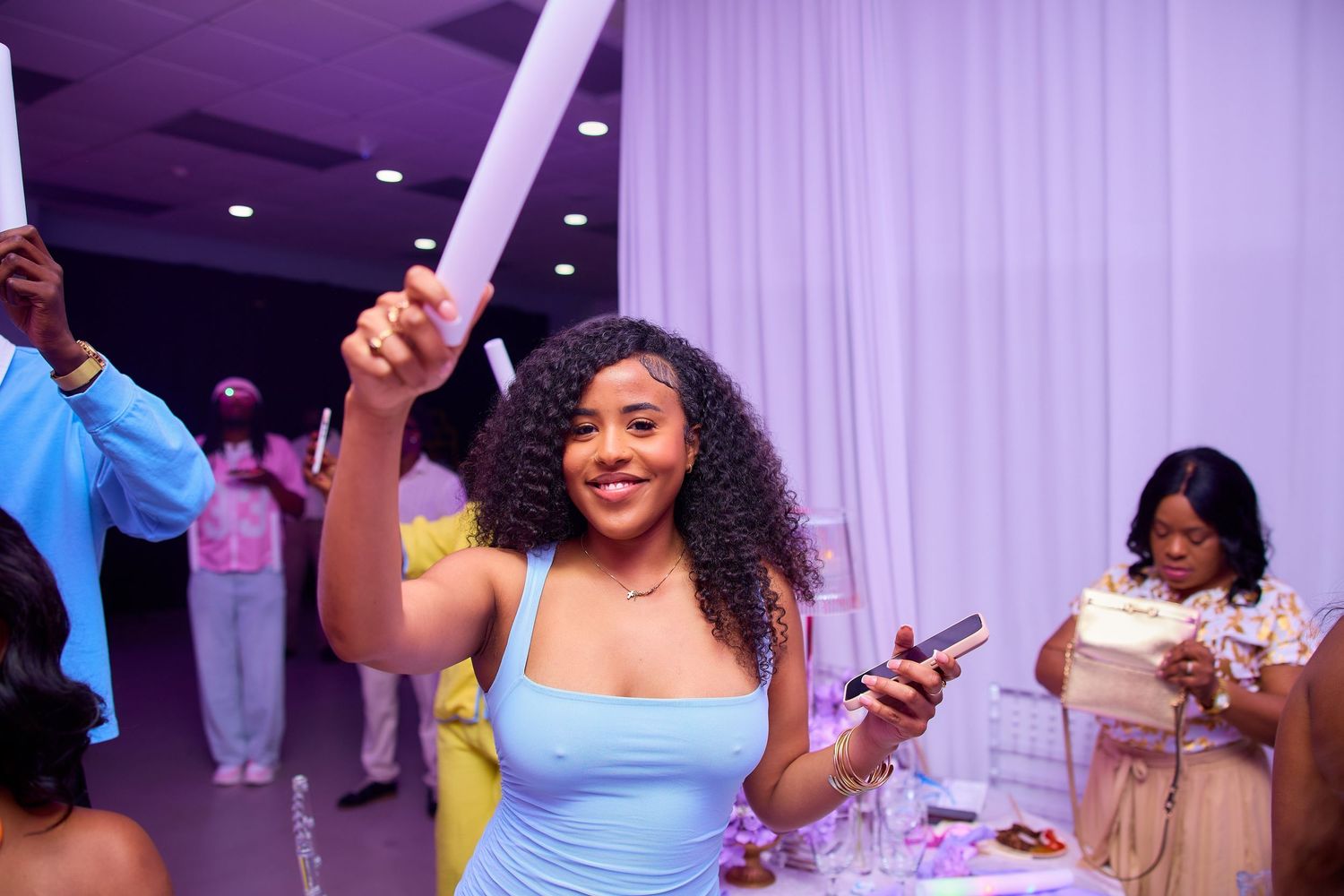 Woman in blue dress dancing and holding a foam stick at an indoor celebration event.