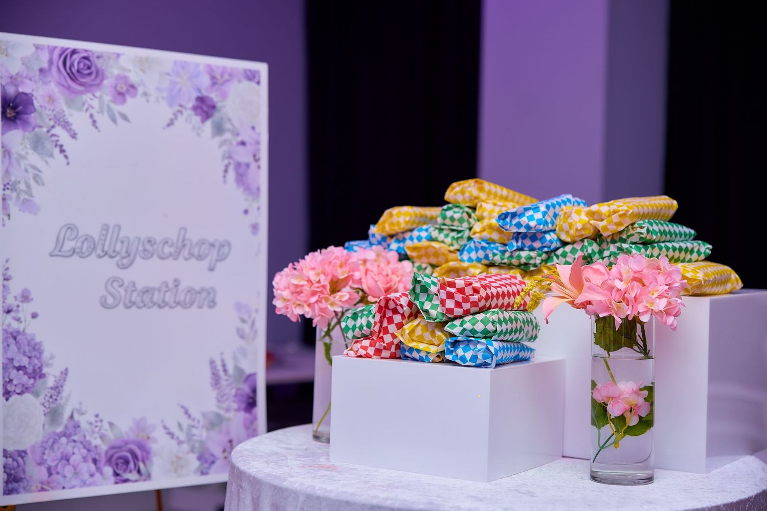 Lollypop station display with colorful wrapped candies and pink flowers at a purple-themed party.